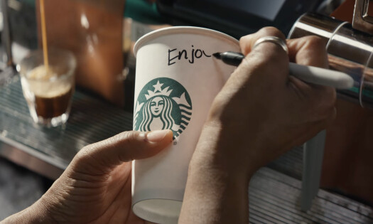 A screenshot from Starbucks' "Magic of Coffee" ad showing a barista's hand writing "Enjoy" on a Starbucks cup