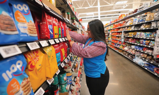 Walmart employee stocking packaged goods on a brightly lit grocery aisle, organizing products on shelves while facing a digital price label system.