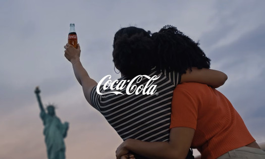 Two girls with their arms around each other, holding a bottle of Coca-Cola with the Statue of Liberty in the background