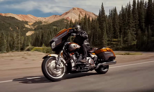 A man rides his orange Harley-Davidson on the road with the mountains as backdrop