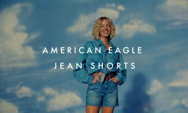 Sydney Sweeney in a denim button down shirt and a pair of denim shorts with a brown belt to tie off the ensemble. She is smiling with her hands in her pocket, standing in front of blue sky and white clouds back drop