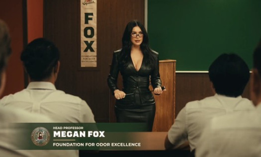 Megan Fox in fitted leather suit, in a lecture hall. A group of students in uniform have their backs to the camera.