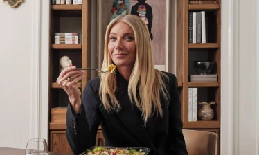 Gwyneth Paltrow is about to take a bite of a goop kitchen salad. A glass of water and salad dressing is on the side.