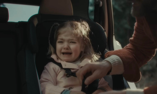 A screenshot from Nissan's "Happy Chaos" ad showing a father buckling his crying daughter into the Rogue 2026
