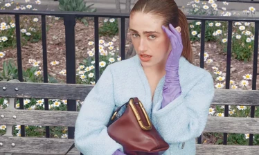 Rachel Sennott sits on a park bench wearing a pastel blue jacket, floral skirt, and purple gloves, holding a burgundy Marc Jacobs bag from the Scene collection.