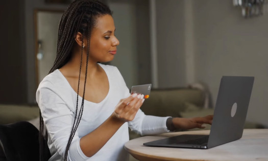 Woman holding a Mastercard while using a laptop, representing agentic commerce and AI-driven payment transactions.