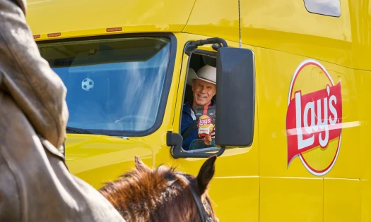 Will Ferrell in the driver's seat of a Lay's-branded yellow truck. He holds a bag of chips out to a person on a horse.