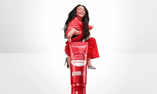 Model in a red outfit reaches toward the camera behind a large Colgate Optic White Pro Series toothpaste tube.