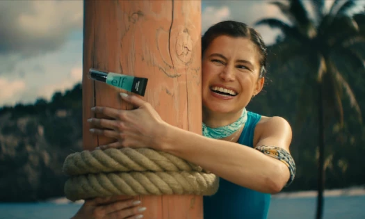 Delaney Rowe smiles while holding onto a wooden challenge post with an e.l.f. Power Grip Primer attached in a Survivor 50 campaign scene.