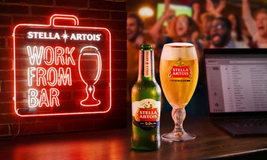 Stella Artois “Work From Bar” campaign visual showing a neon WFB sign, a Stella Artois 0.0 bottle, a chalice, and a laptop on a bar during a soccer match.