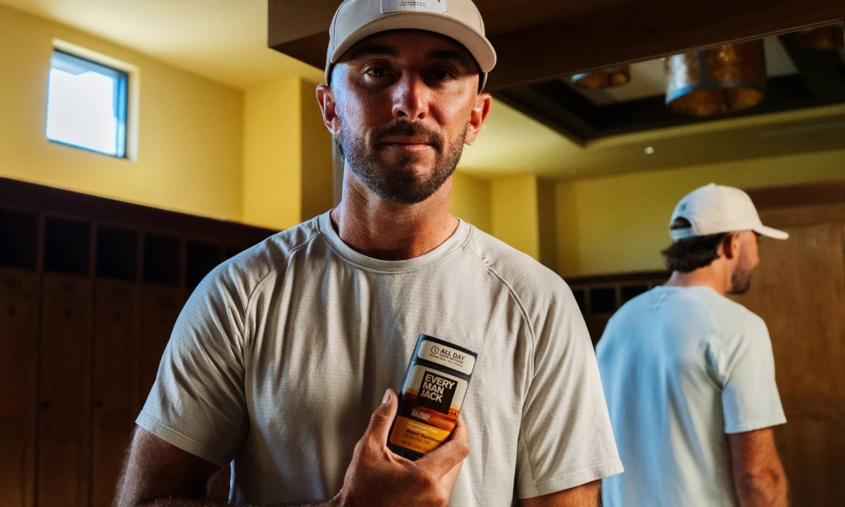 Max Homa holds up an Every Man Jack deodorant to his chest with a mirror reflection in the background
