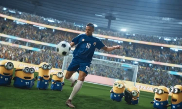 Kylian Mbappé in a blue No. 10 kit kicks a ball on a stadium pitch as Minions in matching uniforms watch.