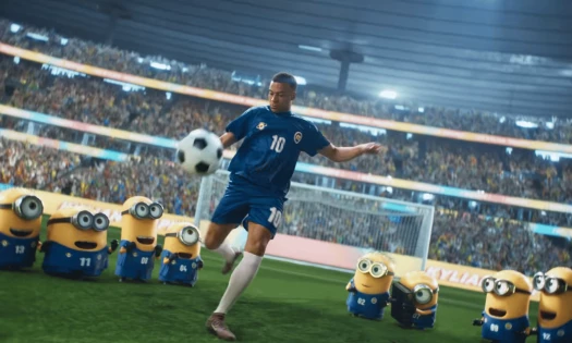 Kylian Mbappé in a blue No. 10 kit kicks a ball on a stadium pitch as Minions in matching uniforms watch.
