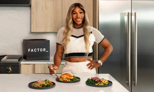 Serena Williams poses with three Factor meals on a kitchen counter and a Factor box on a counter in the background.