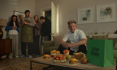 Gordon Ramsey sits on a couch with an Uber Eats delivery and a family keeping their distance in the doorway.