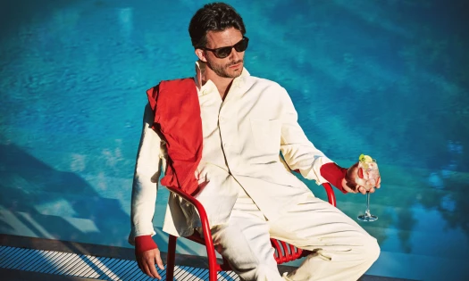 Jonathan Bailey in a cream suit and sunglasses sits by a pool holding a Martini Bianco Spritz.