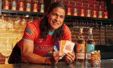 Cristo Fernández in a red Tequila CAZADORES jersey holds colorful “Carded by Cristo” fan cards beside two tequila bottles and a cocktail at a bar.