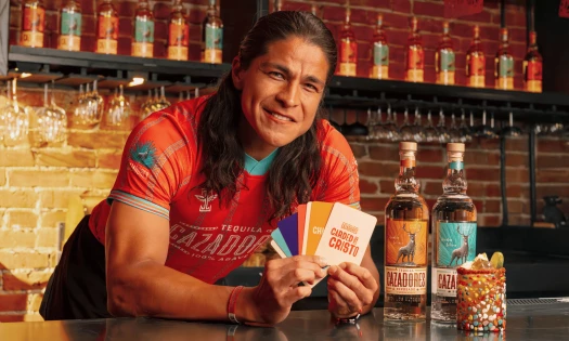 Cristo Fernández in a red Tequila CAZADORES jersey holds colorful “Carded by Cristo” fan cards beside two tequila bottles and a cocktail at a bar.
