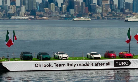 Six Italian cars on a barge on Elliot Bay in Seattle with Italian flags on either side. A banner alongside the barge reads: Oh look, Team Italy made it after all. Turo