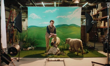 A man poses with two sheep on a staged Vermont landscape inside Darn Tough’s sock factory, surrounded by visible production lights and equipment.