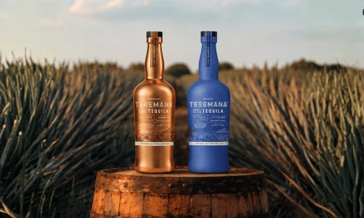 Teremana Reposado and Blanco Craft Edition bottles in metallic copper and blue stand on a wooden barrel in an agave field.