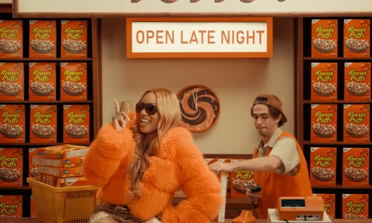 GloRilla in an oversized orange faux-fur coat and sunglasses poses in front of shelves filled with Reese’s Puffs cereal boxes.