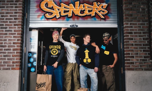 Four men wearing Grindr graphic apparel pose outside a Spencer’s store beneath the retailer’s graffiti-style sign.