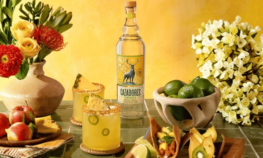 A bottle of CAZADORES Pineapple Infused Tequila stands behind two yellow pineapple cocktails, surrounded by limes, fruit, flowers, and tacos.