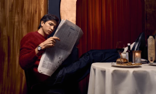 Nicholas Braun wearing a Shinola watch reads a newspaper at a restaurant table with a glass of wine, food, and a candle beside him.