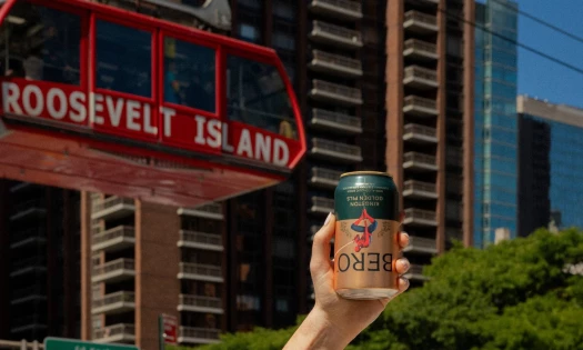 A hand holds up the BERO x Spider-Man: Brand New Day collectible can. A Roosevelt Island cable car hangs in the background.
