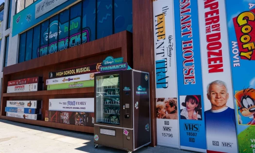 Giant depiction of classic Disney movies in their VHS and DVD media formats. A Disney+ Hulu vending machine sits in front of the billboard.