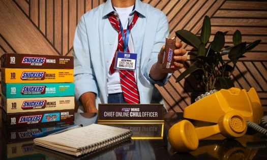 A dishevelled person with the name tag: "Chief Online Chill Officer." SNICKERS Ice Cream Minis boxes are placed on a work desk with the job title plaque, dial telephone, and notepad.