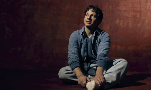 Charlie Puth wearing a blue jacket as he sits on the floor