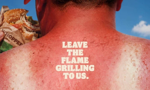 The back of the neck of an aggressively sun burnt man with a shirt outline. He is eating a Burger King Whopper and the tagline: Leave The Flame Grilling To Us" sits the middle of his back.