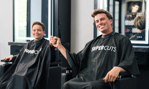 Jackson Olson and a child in Supercuts salon capes, fist pumping.