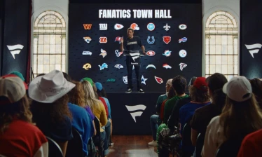 Luke Wilson stands at a podium in a city hall in front of a crowd and a "Fanatics Town Hall" banner with NFL team logos behind him.
