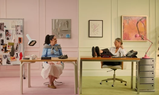 Meg Stalter and Jenna Lyons sitting in parallel desks