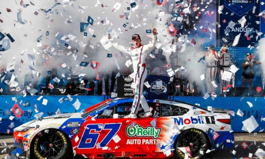 23XI Racing driver Corey Heim standing on the window rim of his no. 67 car sponsored by Mobil 1 and O'Reilly Auto Parts in Victory Lane.