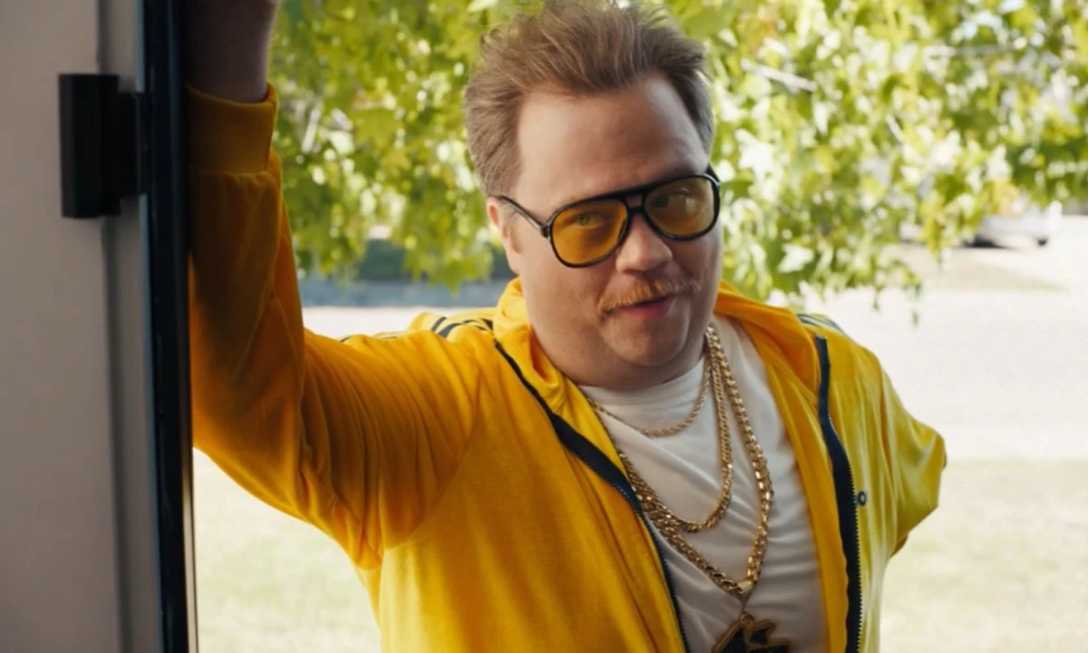 Paul Walter Hauser in a yellow Underdog tracksuit, sunglasses, and gold chains.