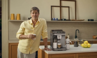Brad Pitt holds a cup of coffee and poses next to the De'Longhi Magnifica Duo.