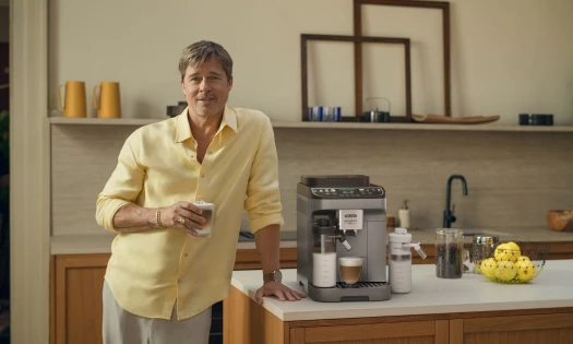 Brad Pitt holds a cup of coffee and poses next to the De'Longhi Magnifica Duo.