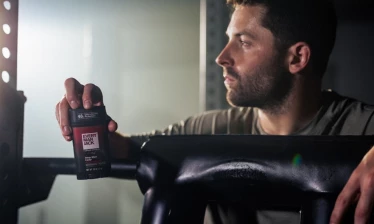 Baker Mayfield leaning on a barbell in a gym holding the Every Man Jack x Baker Mayfield 48-Hour Sweat Control Antiperspirant: Deep Shot Cedar.