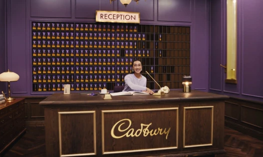 A woman sits behind a Cadbury branded reception desk with chocolate lining the purple wall behind her.