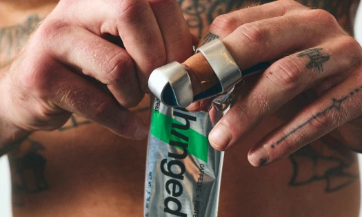 Close-up of tattooed hands opening a silver Unhinged protein and caffeine bar wrapper with green and black branding.
