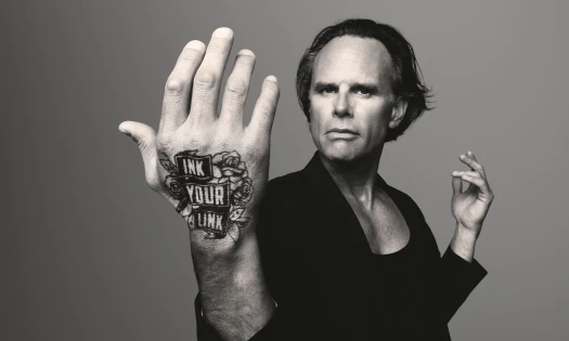 Greyscale photo of Walton Goggins with a "Ink Your Link" tattoo on his hand.