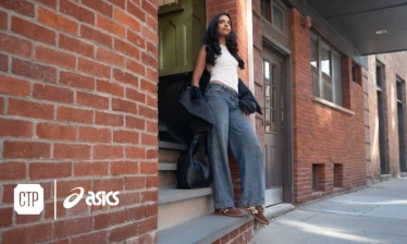 A woman standing at the foot of the steps of a brick-faced building, wearing a pair of ASICS.