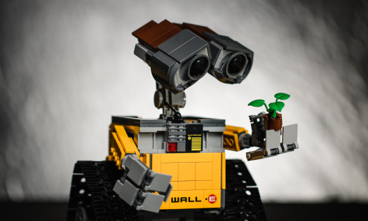 An image of a Lego-built Wall-E robot holding, and looking at, a little Lego plant in front of a dark, grungy background