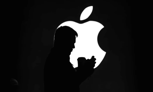Silhouette of a man browsing on his smartphone while standing in front of a lit-up Apple logo