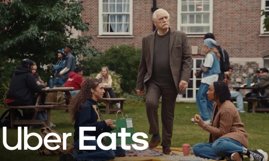 Brian Cox Casually Steps Onto a Picnic Cloth While Walking Between Two Students on a Campus