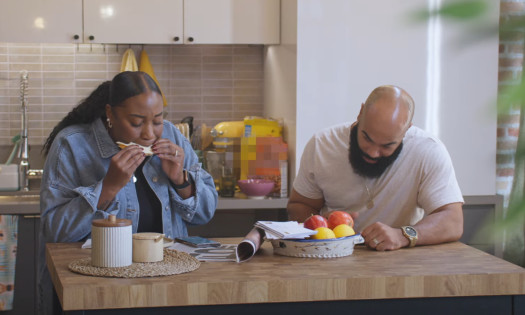 A woman is taking a bite out of Delimex Crispy Quesadillas in the kitchen with a man beside her.